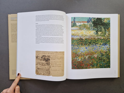 An open book laid flat on a dark gray surface, showing printed text on the left page and a colorful impressionist-style landscape painting on the right page. A person's finger is visible holding the left edge of the book.