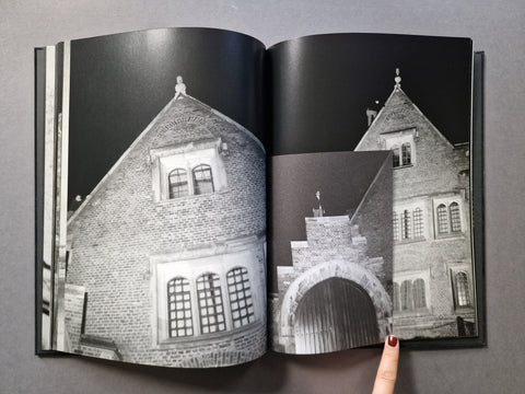 An open hardcover book showing black-and-white architectural photographs of brick buildings and a stone archway, with a finger pointing at the right page, on a gray surface.