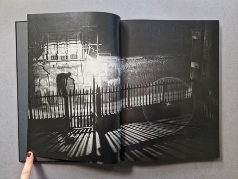 An open hardcover book displaying a high-contrast black and white photographic print of a person standing behind a fence in a moody urban setting.
