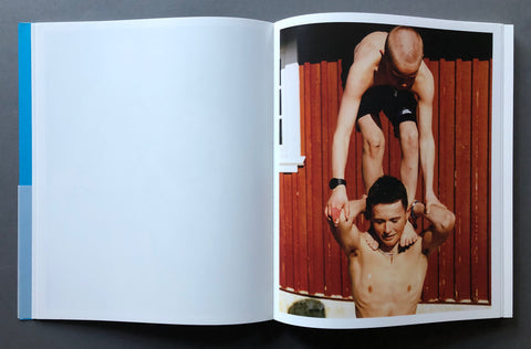 An open book or magazine laid flat on a dark surface, showing a large blank white page on the left and a printed photograph on the right with two shirtless men posed against a red wooden background.