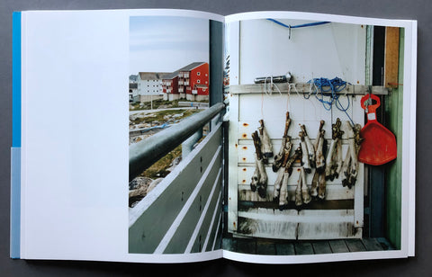 An open spread of the book to-skip-a-sinking-stone by daniel-dorsa laid flat on a dark gray surface, showing two printed photographs side by side. The left page features a photograph of a coastal walkway with red and white buildings in the distance.
