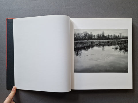 An open book - The right page features a black-and-white landscape photograph of a calm lake or river with bare trees along the far bank and ripples on the water.
