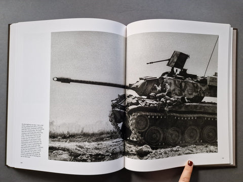 An open book showing a black-and-white photograph of a tank on a battlefield, photographed from above on a dark gray surface. 