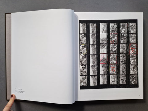An open art book, showing a large white left page with small text and a black-and-white illustrated right page featuring a grid of vertical panels with black, white, and red markings.