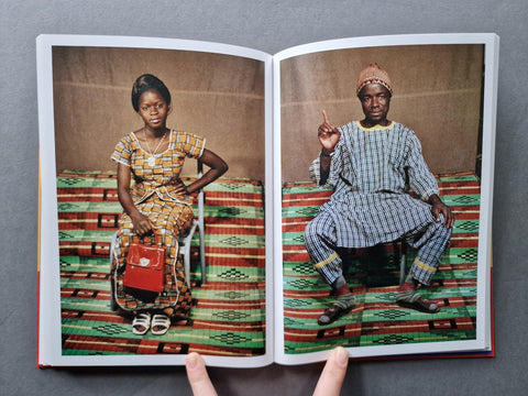 An open book or photo album showing two printed portraits of African people seated on patterned rugs, photographed against a dark gray background with two fingers holding the open pages.