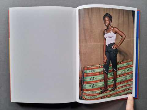 An open book or magazine spread on a dark gray surface, showing a printed fashion photograph on the right page of a person standing on a patterned green and red bench against a tan backdrop.