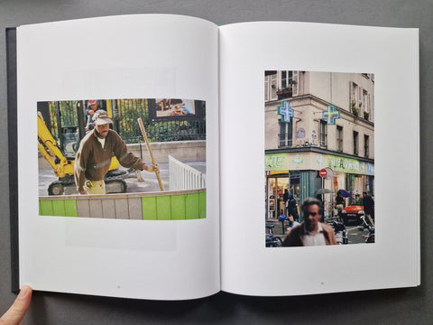An open art book. The left page features a color photograph of a construction worker near a yellow excavator, and the right page shows a street scene with a pharmacy and pedestrians.