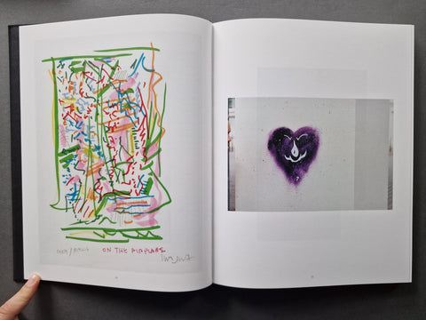 An open art book showing two printed pages. The left page has a colorful abstract line drawing with green, pink, orange, and blue scribbles, and the right page shows a square photo insert with a purple heart-shaped mark on a light background.