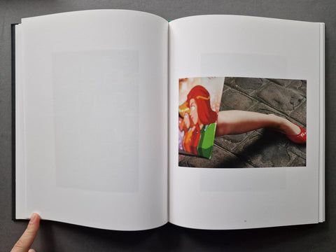 An open hardcover book photographed from above on a dark gray surface. The left page is blank, and the right page shows a small printed photograph of a person's leg in a red shoe against a stone floor, with a finger holding the left page open.