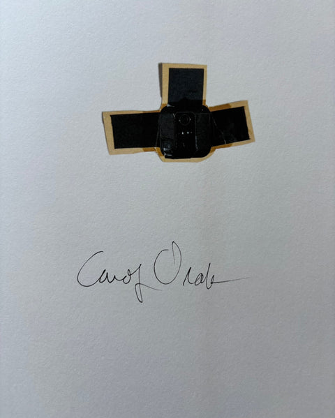 A small black cross-shaped adhesive patch with gold edges is placed on a white textured surface, with a cursive signature printed below it.