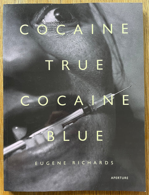 The photobook cover of  Cocaine True cocaine Blue by Eugene Richards. In dsoftcover black and white with a photo of a woman holding a needle between her teeth.
