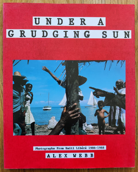 The photography book of Under a Grudging Sun: Photographs from Haiti Libere 1986-1988 by Alex Webb. In softcover red.