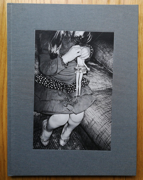 The photobook cover of To Belong by Anders Petersen. Hardback in grey with image of a girl playing with a doll.
