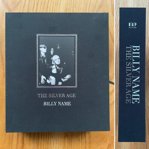 The photobook cover of The Silver Age by Billy Name. Hardback in navy blue with a central image of Andy Warhol with 3 women. Signed by multiple people.