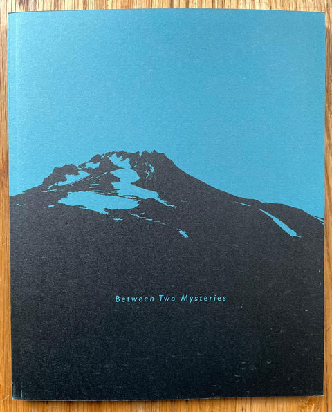 The photography book cover of Between Two Mysteries by Carl Bigmore. Paperback in blue with mountain in black.