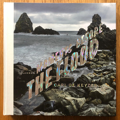 The photography book cover of Moments before the Flood by Carl de Keyzer. Hardback with image of the sea and rocks on the cover, title is metallic/colourful.