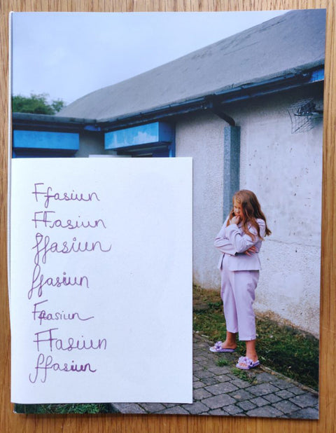 The photography book cover of Ffasiwn by Clementine Schneidermann. Paperback with a girl standing in lilac clothes. "Ffasiwn" written in 7 different styles.