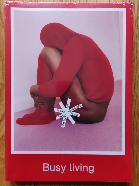 The photography book cover of Busy Living by Coco Capitan. Paperback in red with a person sat in a red hoodie and socks with their arms wrapped around their knees.