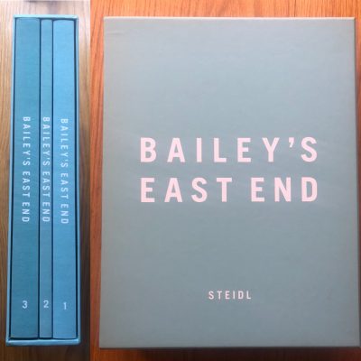 The photography book cover of Bailey's East End by David Bailey. Hardback grey cover with 3 blue books.