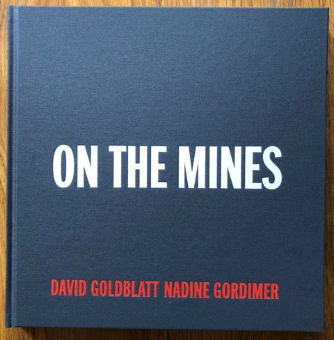 The photography book cover of On the Mines by David Goldblatt and Nadine Gordimer. Hardback in blue with white and red text.