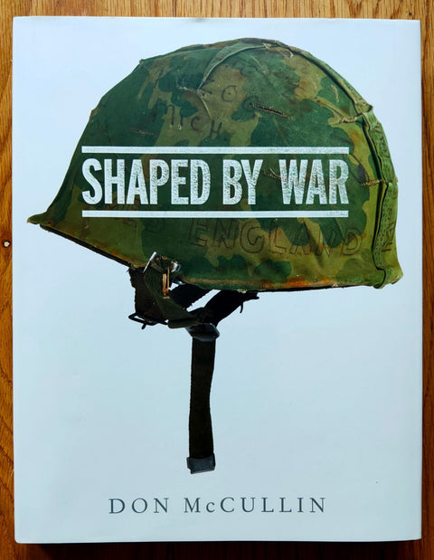 The photography book cover of Shaped by War by Don McCullin. Hardback in white with a camouflage combat helmet on the cover. Signed.