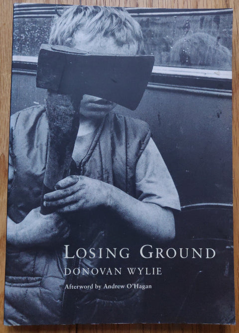 The photography book cover of Losing Ground by Donovan Wylie. Paperback in B&W with image of a boy holding an axe in front of his eyes.