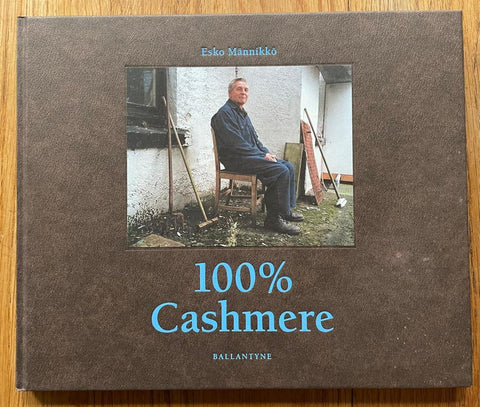 The photography book cover of 100% Cashmere by Esko Manniko. Hardback in brown with blue title and image of a man sitting on chair outside.