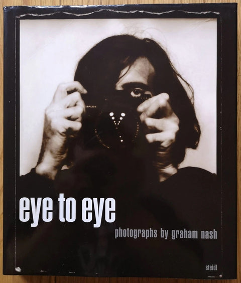 The photography book cover of Eye to Eye by Graham Nash. Hardback with cover photo of a man holding a camera up, in sepia.