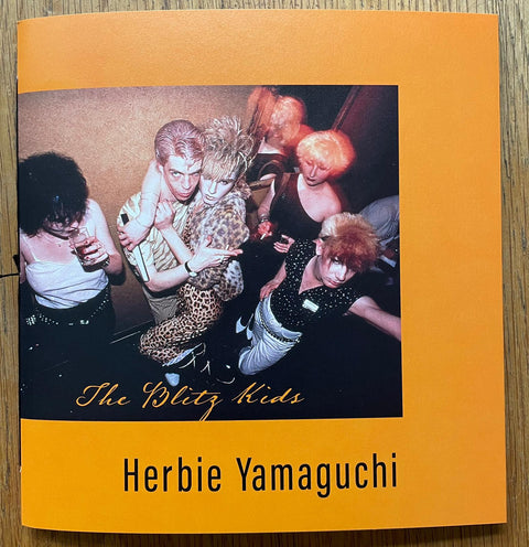 The photography book cover of The Blitz Kids by Herbie Yamaguchi. Paperback in orange with image of a group of dressed up people from above. Signed.