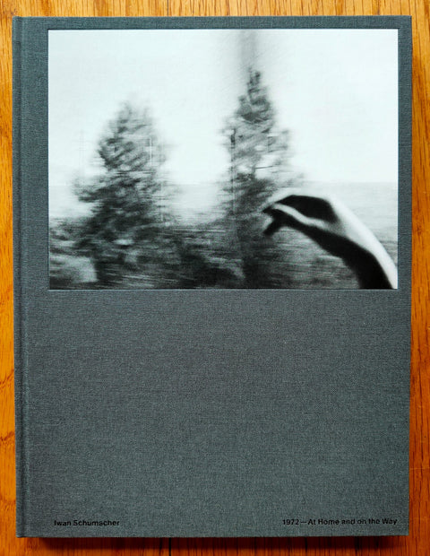 The photography book cover of At Home and on the Way by Iwan Schumacher. Hardback in grey with image of a hand in front of trees.