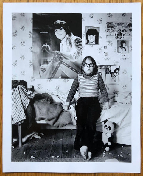 The limited edition print from the photobook The Portraits by John Myers. B&W print of a girl with glasses sitting on her bed.
