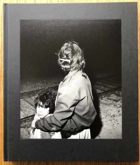 The photography book cover of Midnight La Frontera by Ken Light. Hardback in black with image of a mother holding a child by train tracks.