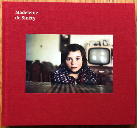 The photography book cover of Un Village / A Village by Madeleine de Sinety. Hardback in red with image of a girl looking into the camera.