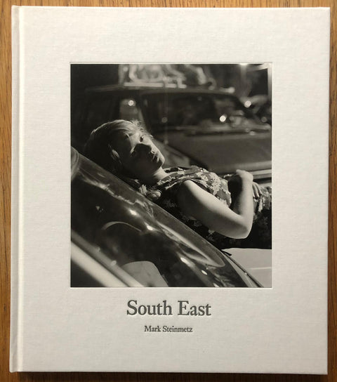 The photography book cover of South East by Mark Steinmetz. Hardback in white with B&W image of a woman lying on the front of a car.