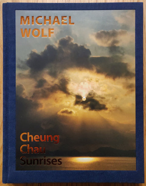 The photography book cover of Cheung Chau Sunrises by Michael Wolf. Hardback with dark blue border and image of the sun peeking through some clouds.