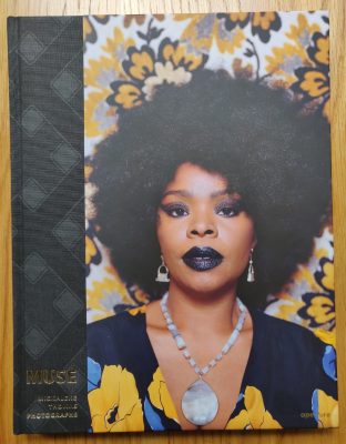 The photography book cover of Muse by Mickalene Thomas. Hardback with a cover image of a woman. Blue and yellow flowers are present both on the woman's dress and the wallpaper.
