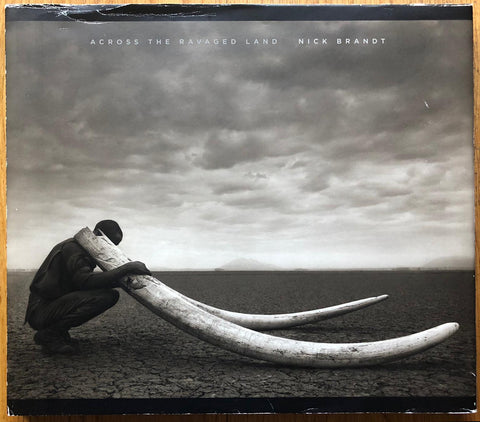 The photography book cover of Across the Ravaged Land by Nick Brandt. Hardback cover with image of a man holding two large tusks. Signed.