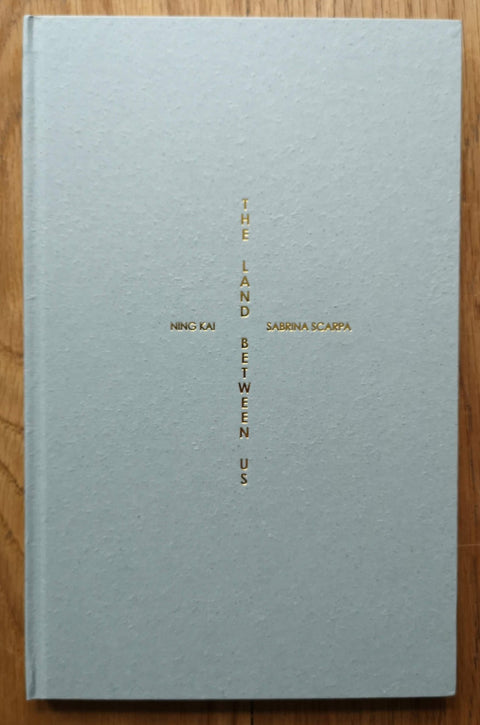 The photography book cover of The Land Between Us by Ning Kai and Sabrina Scarpa. Hardback in white with gold text in a cross shape. Signed in gold pen.