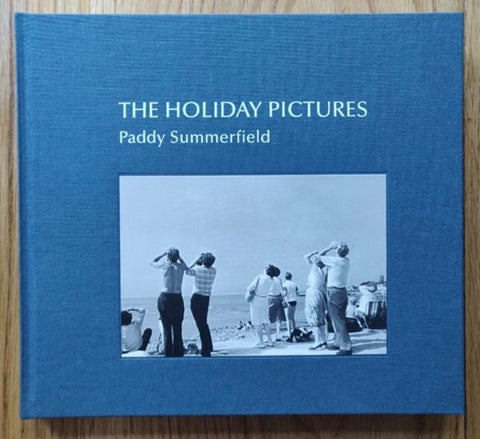 The photography book cover of The Holiday Pictures by Paddy Summerfield. Hardback in blue with image of people standing on the beach looking up to something in the sky.
