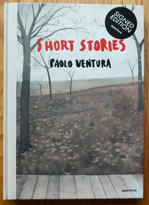The photography book cover of Short Stories by Paolo Ventura. Hardback with cover image of a field with trees as a backdrop behind decking. Signed.