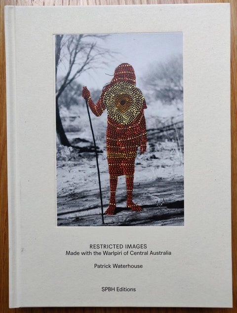 The photography book cover of Restricted Images by Patrick Waterhouse. Hardback in white. B&W image of a person standing holding a stick with Warlpiri dot painting over the top in red and yellow.