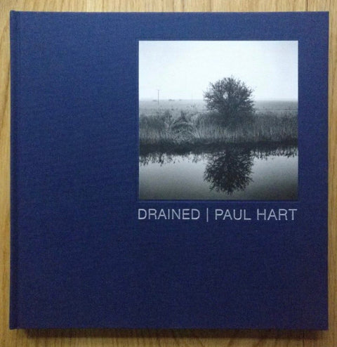 The photography book cover of Drained by Paul Hart. Hardback in dark blue with image of fallen tree in the top right corner.