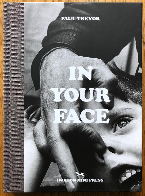 The photography book cover of In Your Face by Paul Trevor. Hardback in B&W with image of a man holding a young boys nose.