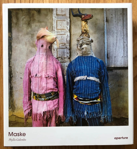 The photography book cover of Maske by Phyllis Galembo. Hardback with one person in pink and one in blue both standing with masks.