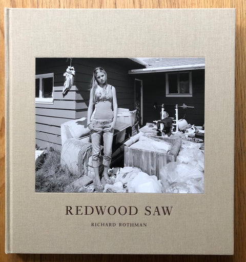 The photography book cover of Redwood Saw by Richard Rothman. Hardback in beige with B&W image of a girl standing by belongings outside a house.