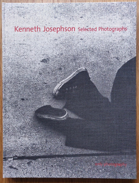 The photography book cover of Selected Photographs by Kenneth Josephson. Hardback B&W image of someone laying on the concrete floor in converse shoes with red text.