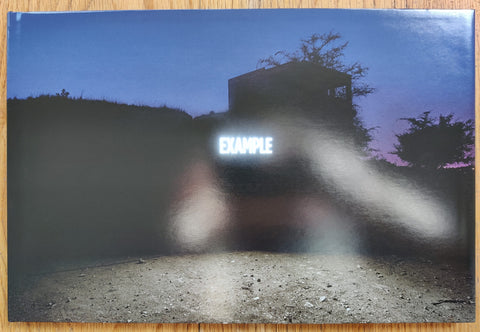 The photography book cover of Facts on the Ground by Shimon Attie. Hardback image of a building in the dark with "EXAMPLE" bright lights.