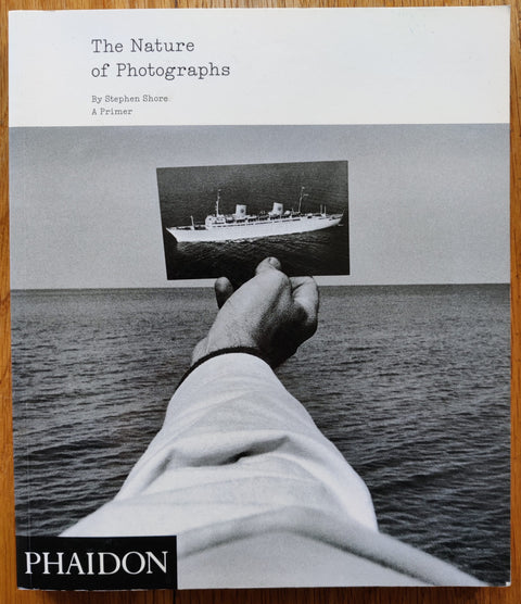 The photography book cover of The Nature of Photographs by Stephen Shore. Paperback in black and white with image of someone holding a picture of a boat in front of the sea.