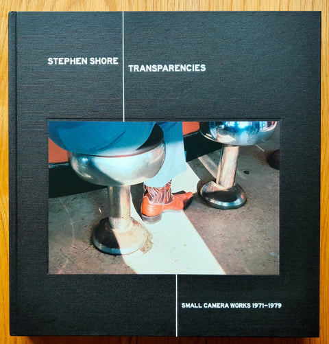 The photobook cover of Transparencies: Small Camera Works 1971-1979 by Stephen Shores. Hardback in black with image of someone sitting on a stool with a red cowboy boot. Signed.