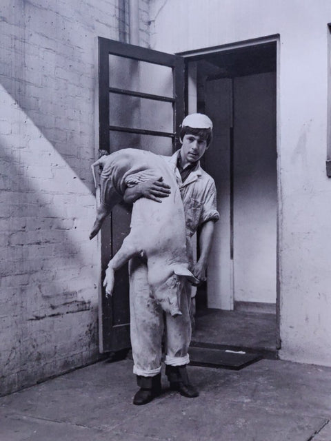 A black and white photograph showing a man in a work uniform standing in a doorway and holding a skinned animal body, likely a pig, in a workshop or studio setting.